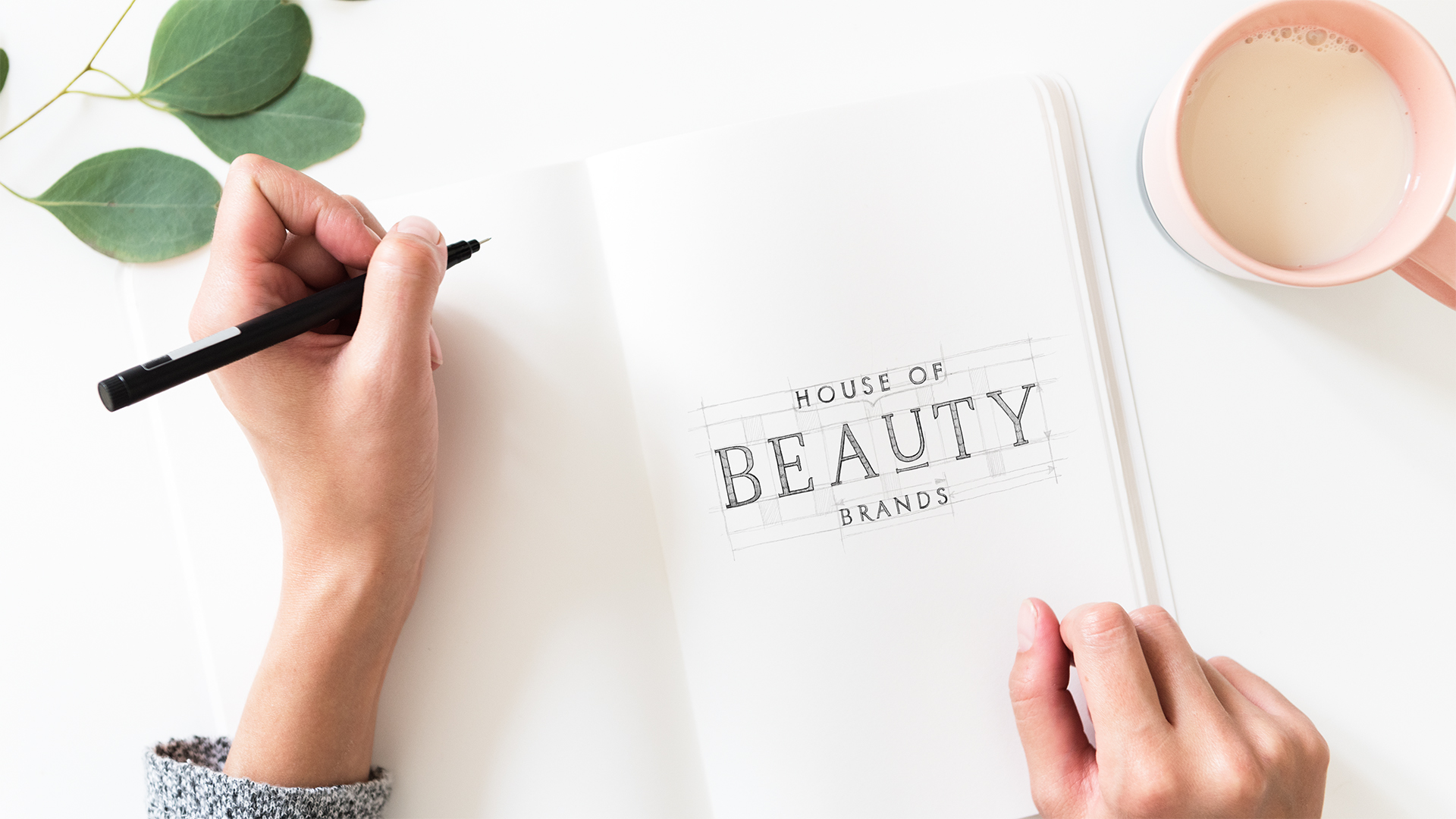 House Of Beauty Brands - Logo naszkicowane na kartce z liniami pomocniczymi w trakcie projektowania, obok ręka z ołówkiem i kawa